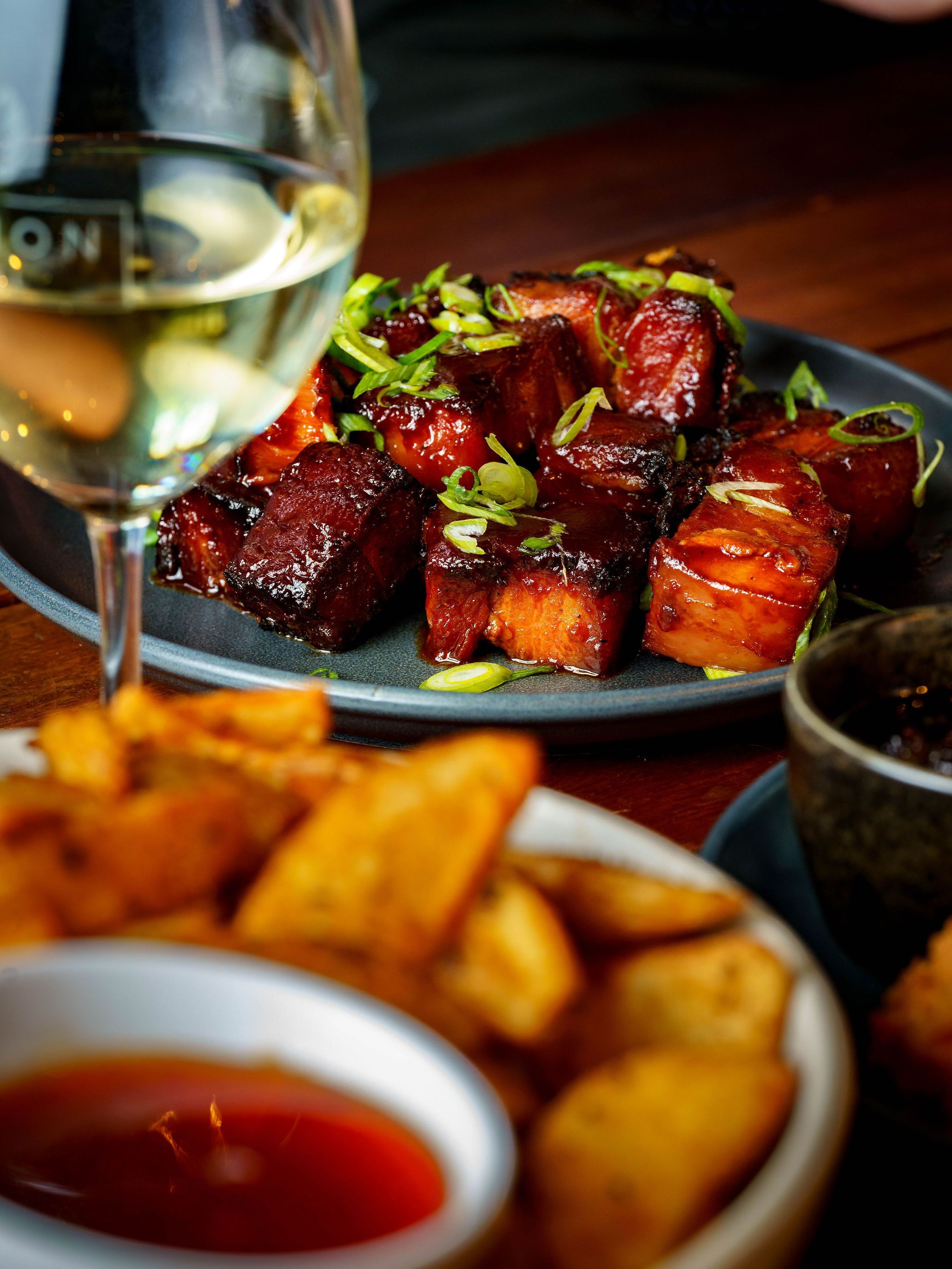 Un plato de carne glaseada con guarnición, acompañado de papas fritas y una copa de vino blanco.