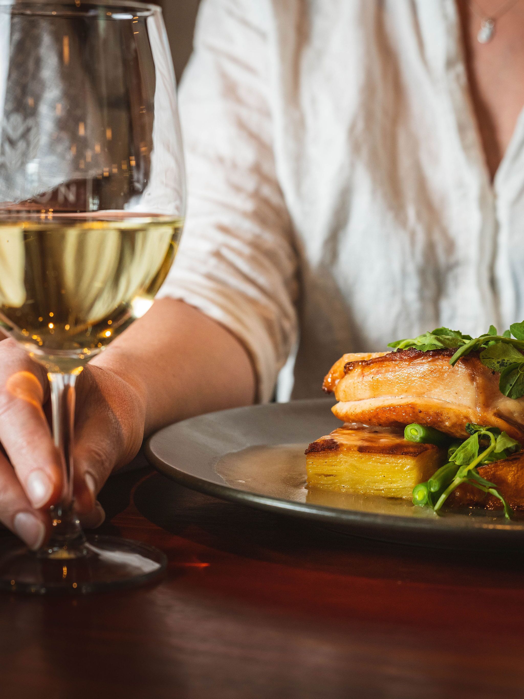 Une personne tenant un verre de vin blanc, assise à une table devant une assiette de nourriture.