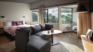 Una habitación de hotel moderna con una cama grande, un sofá gris, una mesita redonda y un balcón con una silla.