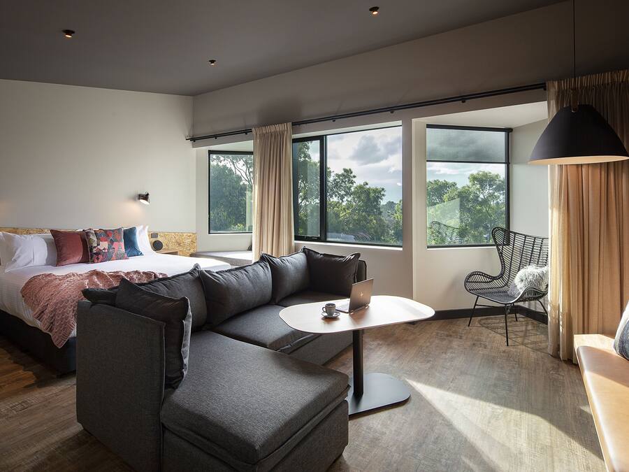 A modern hotel room with a large bed, a gray sofa, a small round table, and a balcony with a chair.