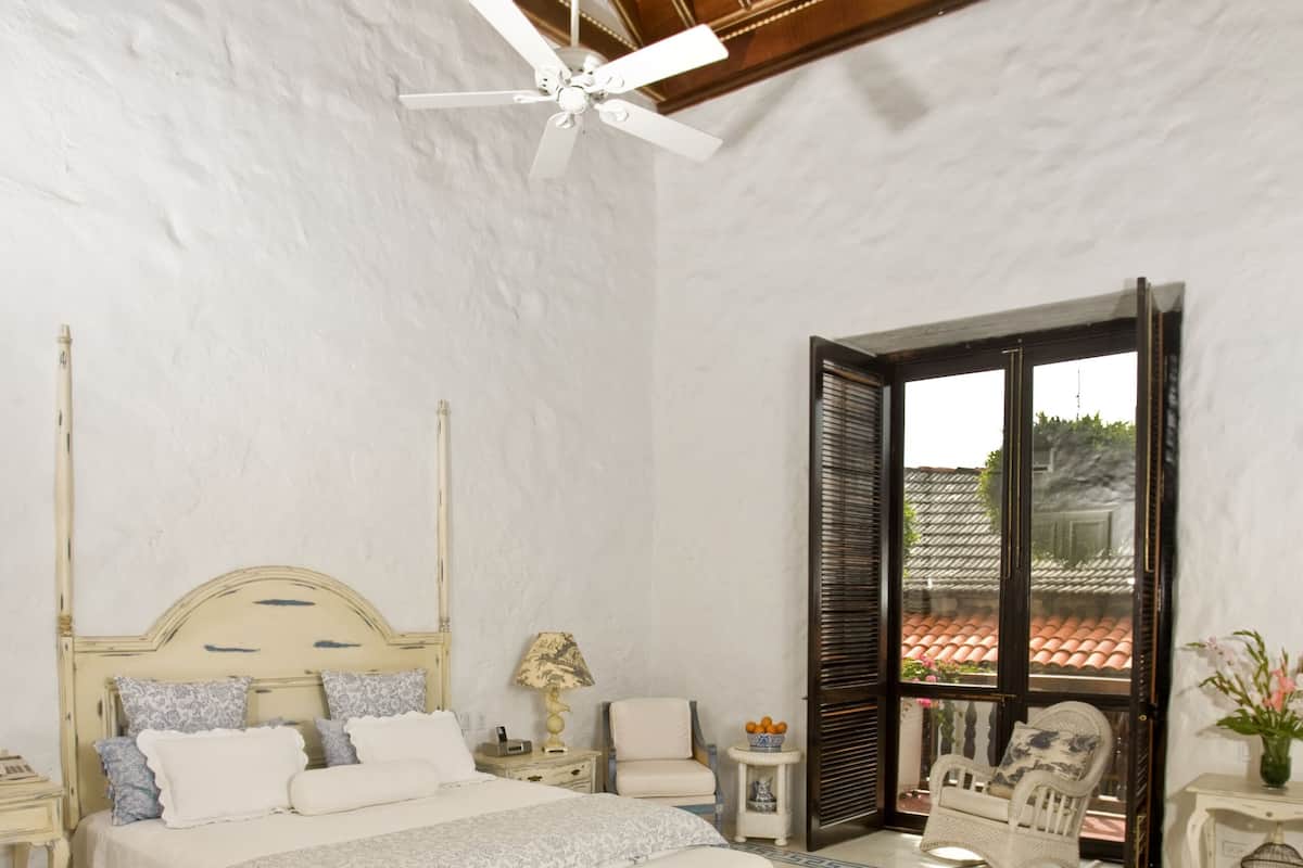 Une chambre avec un grand lit, un ventilateur de plafond, une fenêtre avec des volets et une vue sur l’extérieur.
