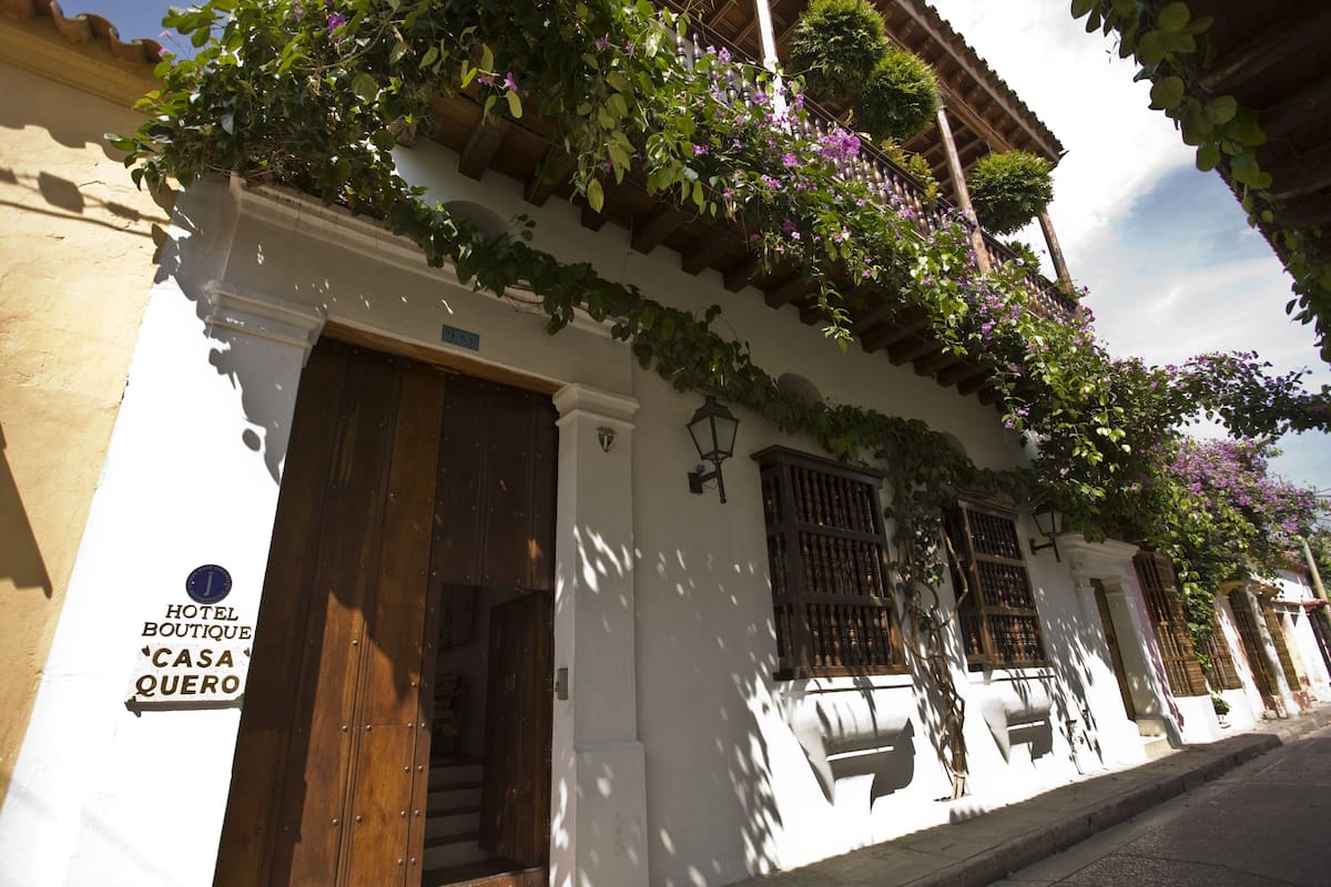 Un bâtiment blanc avec des portes et des fenêtres en bois, un balcon orné de plantes suspendues et une enseigne indiquant qu’il s’agit d’un hôtel.