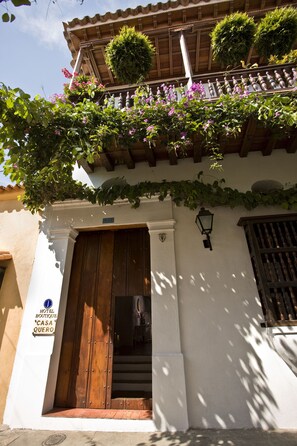 A hotel entrance with a wooden door, a sign reading "HOTEL BOUTIQUE CASA QUERO", and a balcony with plants and flowers. - Casa Quero Hotel Boutique (Cartagena)