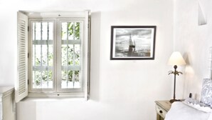 Ein Schlafzimmer mit einem Bett, einem Deckenventilator, einem Fenster mit Fensterläden, einem gerahmten Bild an der Wand und einem Nachttisch mit einer Lampe.
