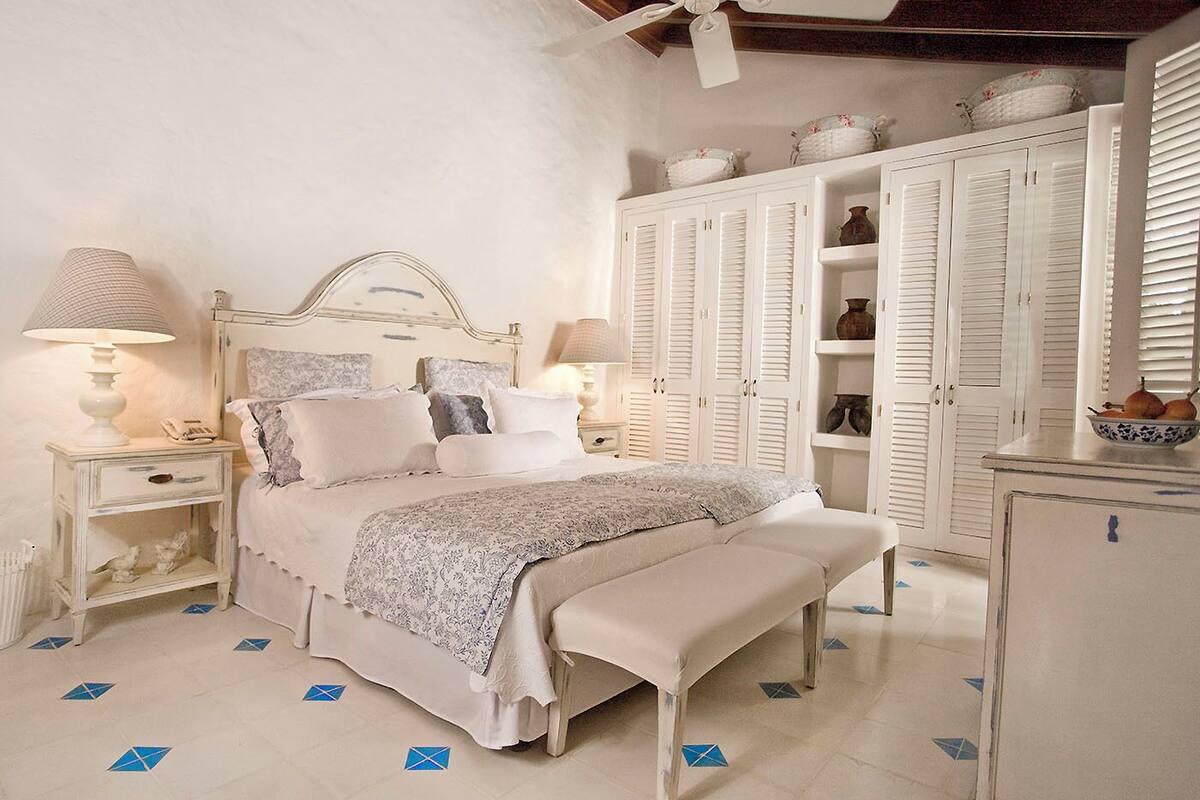 Une chambre à coucher comprenant un lit, des tables de chevet, un banc, une armoire et un ventilateur de plafond.