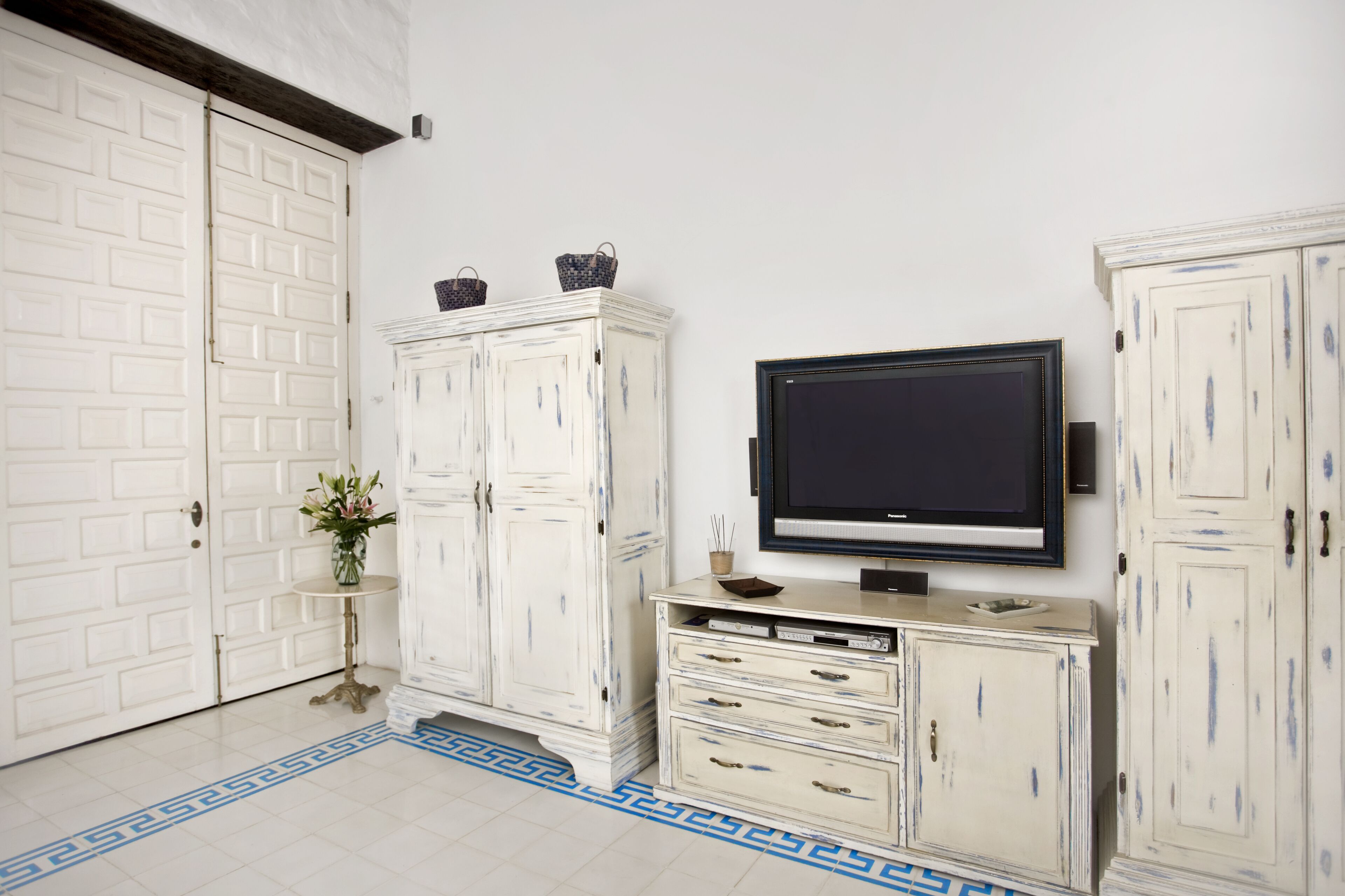 A room with a white door, a white cabinet with two baskets on top, a flat-screen TV on a stand, and a small table with a vase.