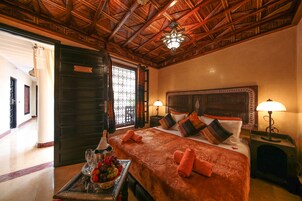 Habitación de hotel con techo de madera, una cama con varias almohadas, una mesita de noche con lámpara y una bandeja con fruta y vasos.