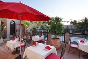 An outdoor dining area with tables and chairs, red umbrellas, and potted plants. - Riad Africa (Marrakech)