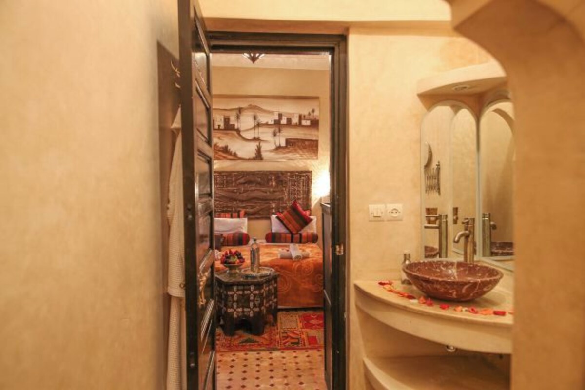 A narrow hallway leading to a bathroom with a large mirror, a sink with a decorative bowl, and a bed with a patterned bedspread.