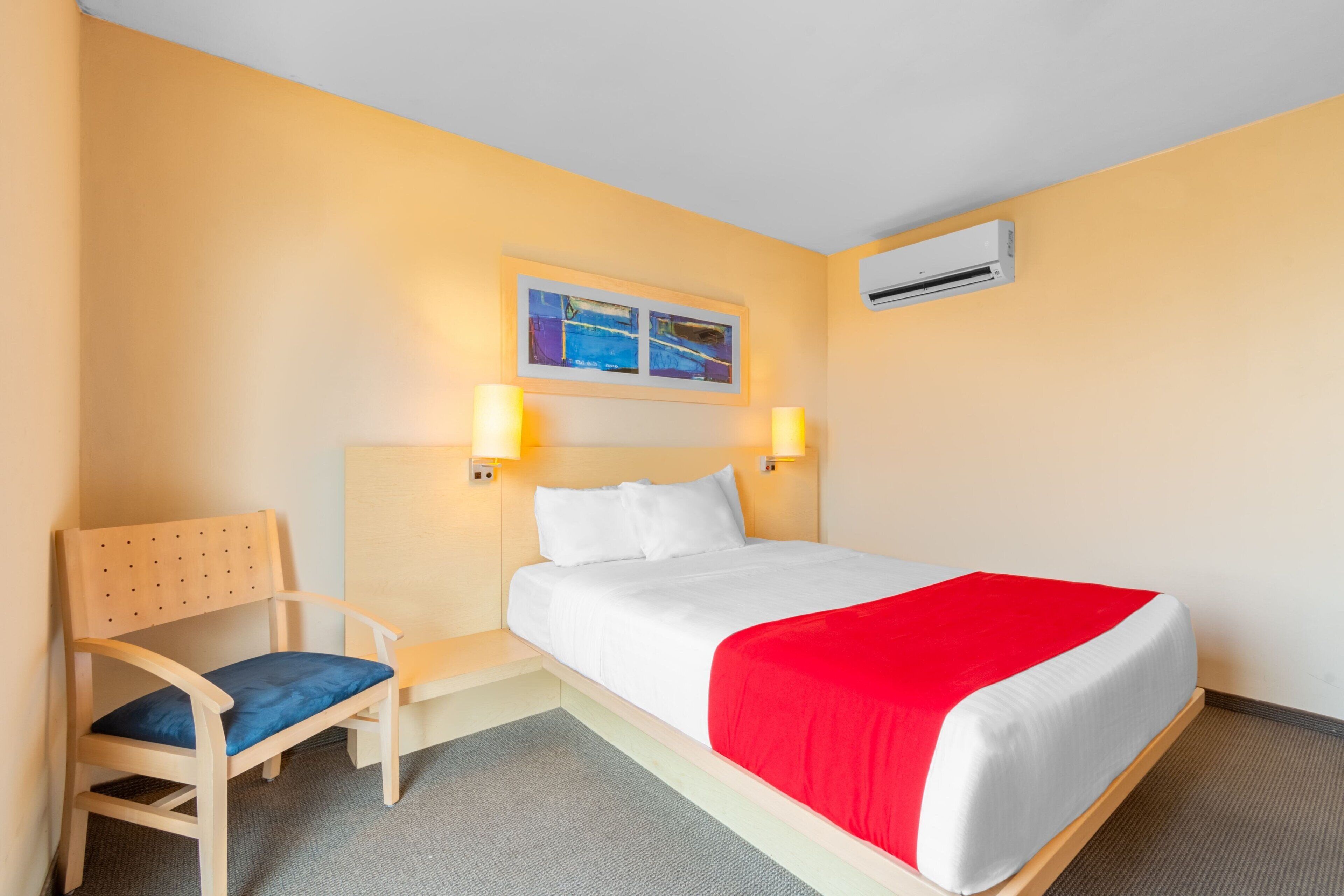 Una habitación de hotel con cama, una silla, una mesita de noche y un equipo de aire acondicionado.