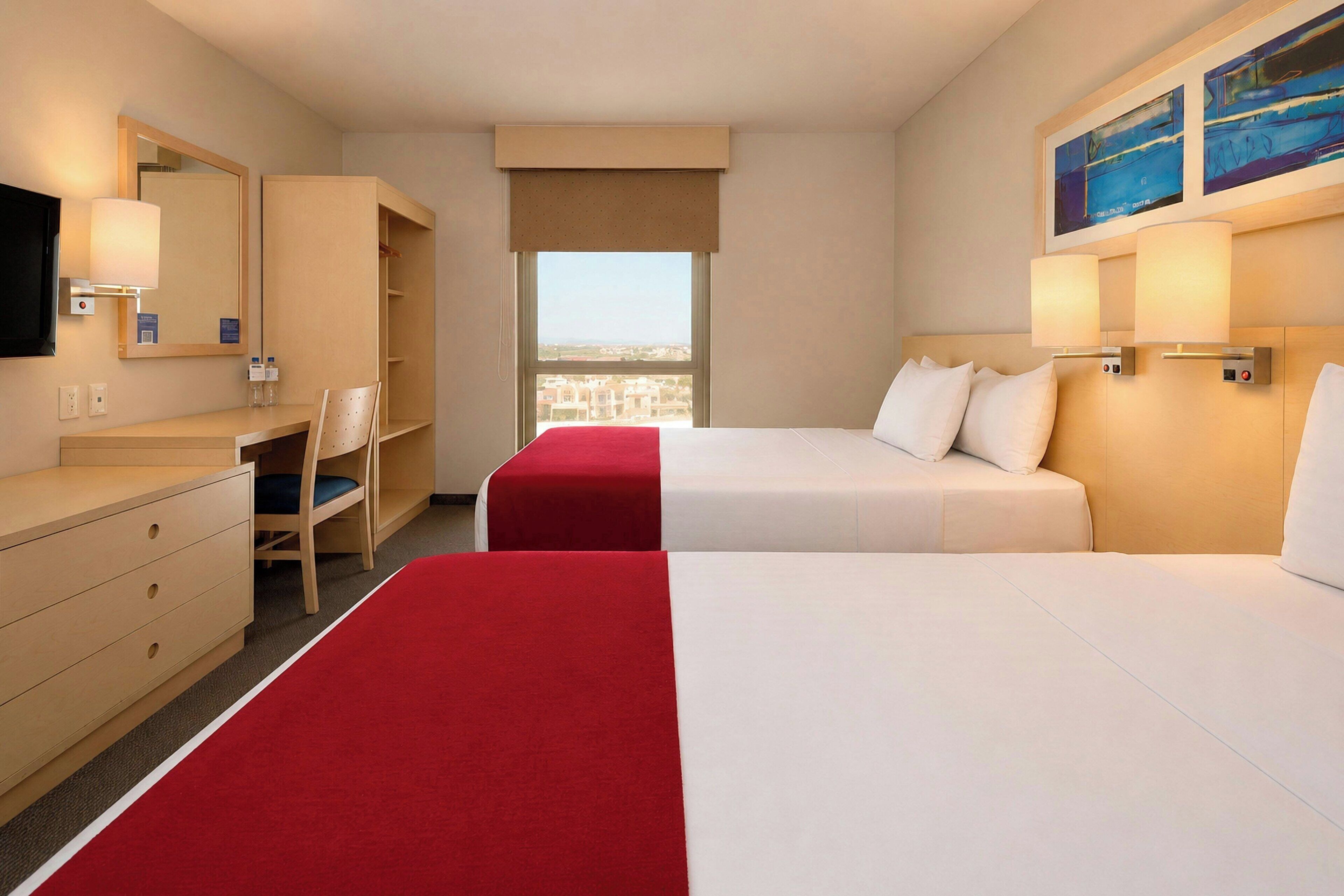 A hotel room with two beds, a desk, a chair, a mirror, and a window with a city view.