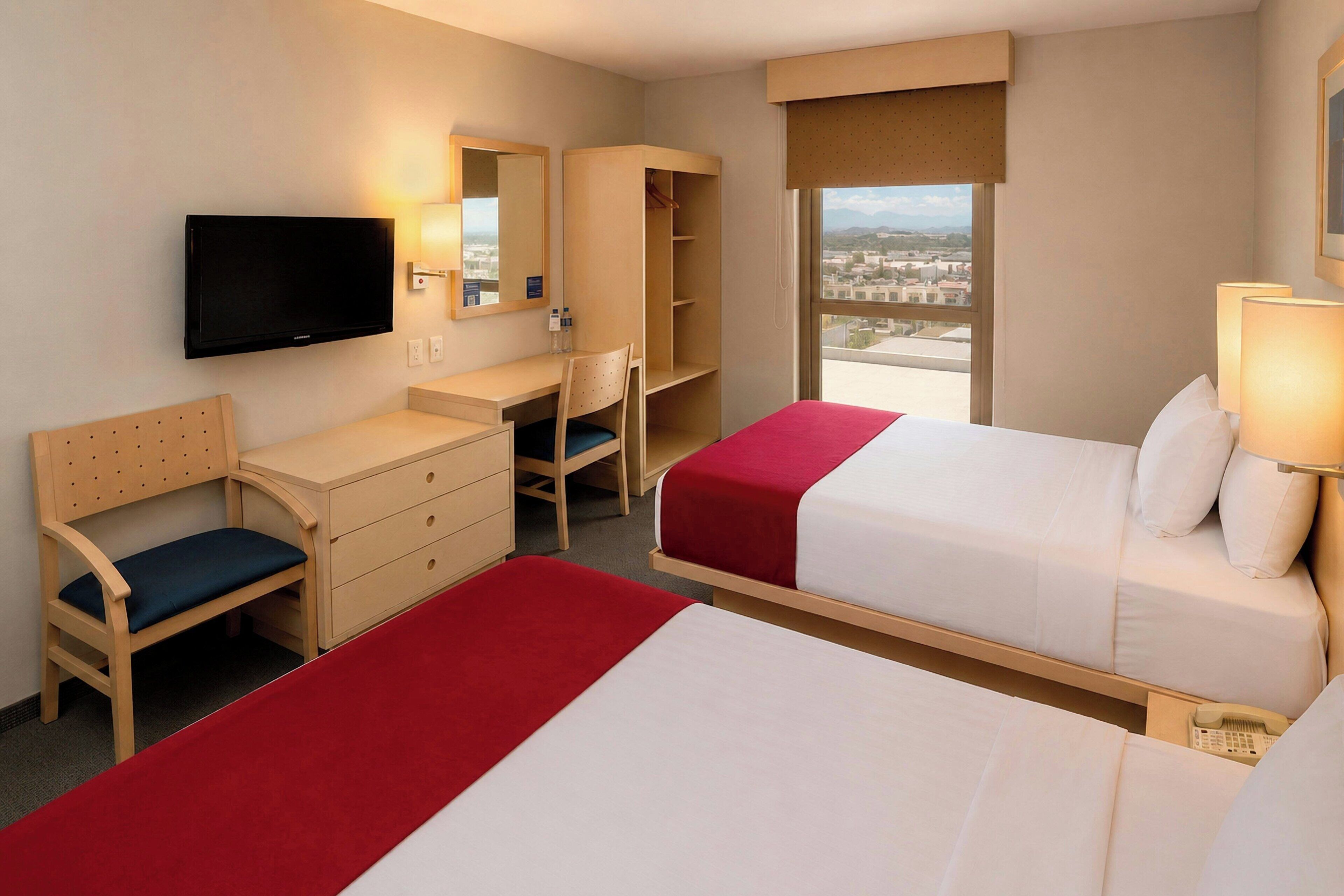 Une chambre d’hôtel avec deux lits, un bureau, une chaise, une télévision, une armoire et une fenêtre donnant sur la ville.