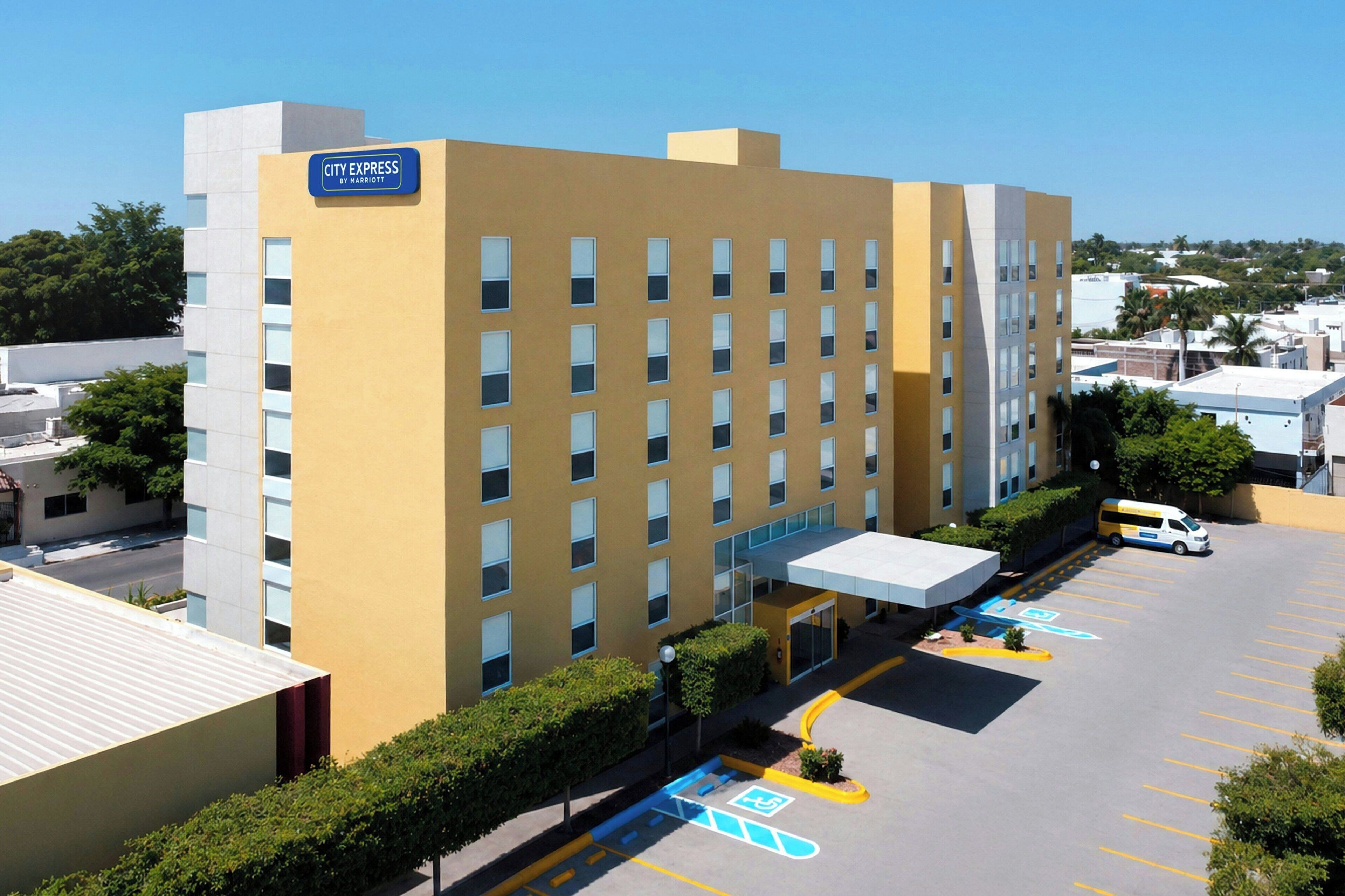 Un hotel de varios pisos con fachada amarilla, un acceso cubierto y un estacionamiento con plazas reservadas para discapacitados.