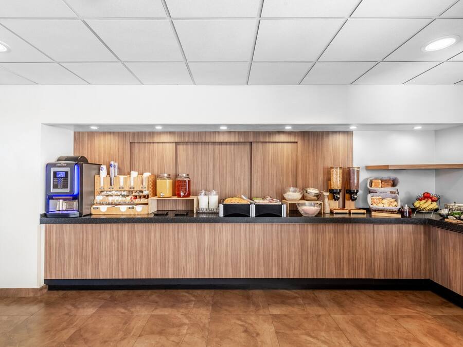 Een modern ontbijtbuffet met een houten achtergrond, voorzien van een koffieapparaat, diverse etenswaren en toevoegingen.