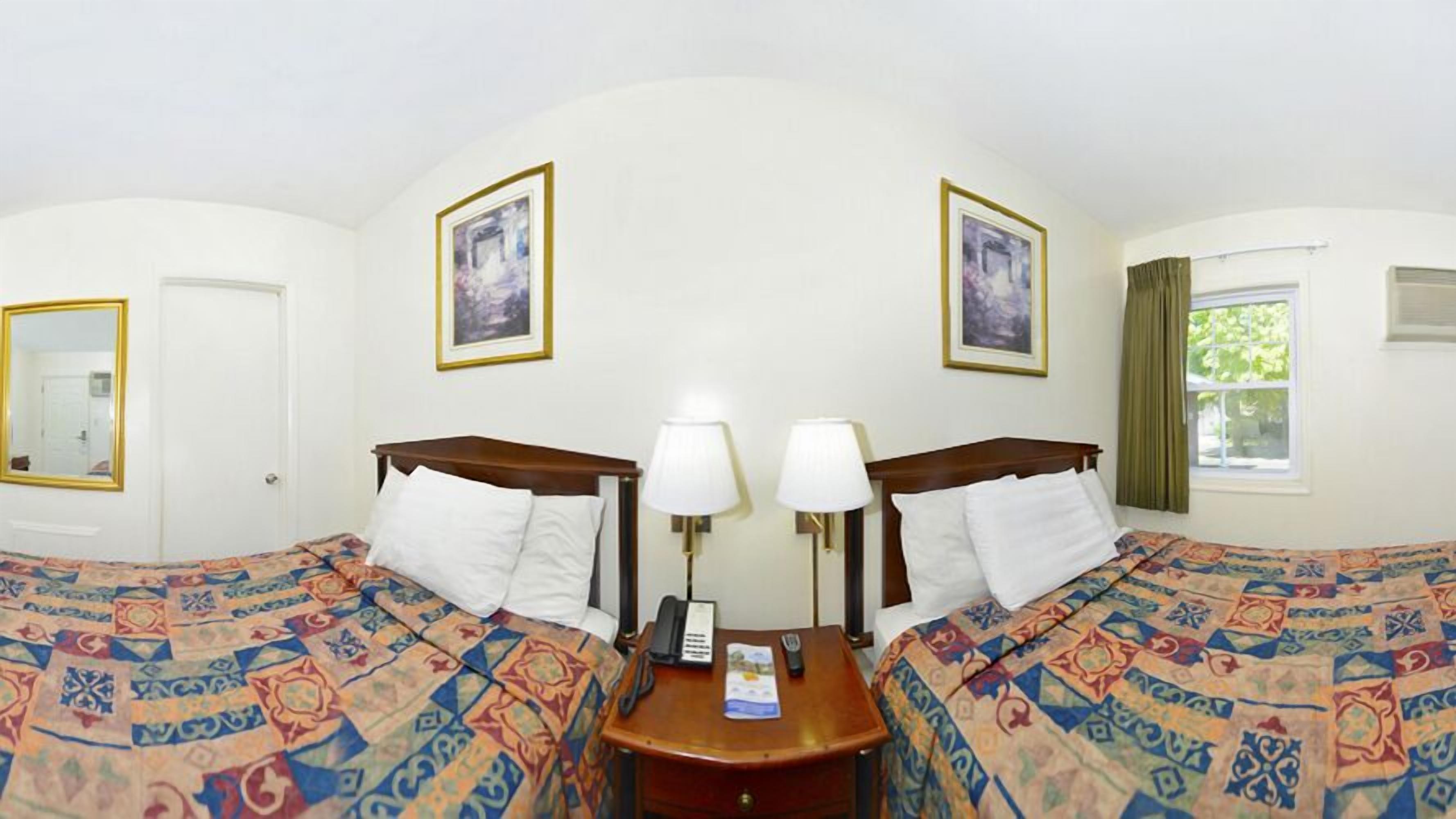 A hotel room with two beds, each with a patterned bedspread, a bedside table with a lamp, and a mirror. There are framed pictures on the wall and a window with curtains.