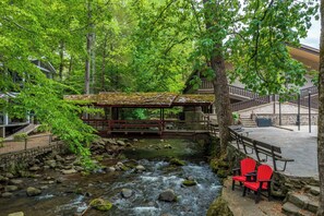 A riverside walkway with benches and a covered area, surrounded by lush greenery and a stone building. - Brookside Lodge (Gatlinburg)