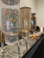 Buffet com três grandes recipientes cheios de cereais coloridos e outros petiscos.