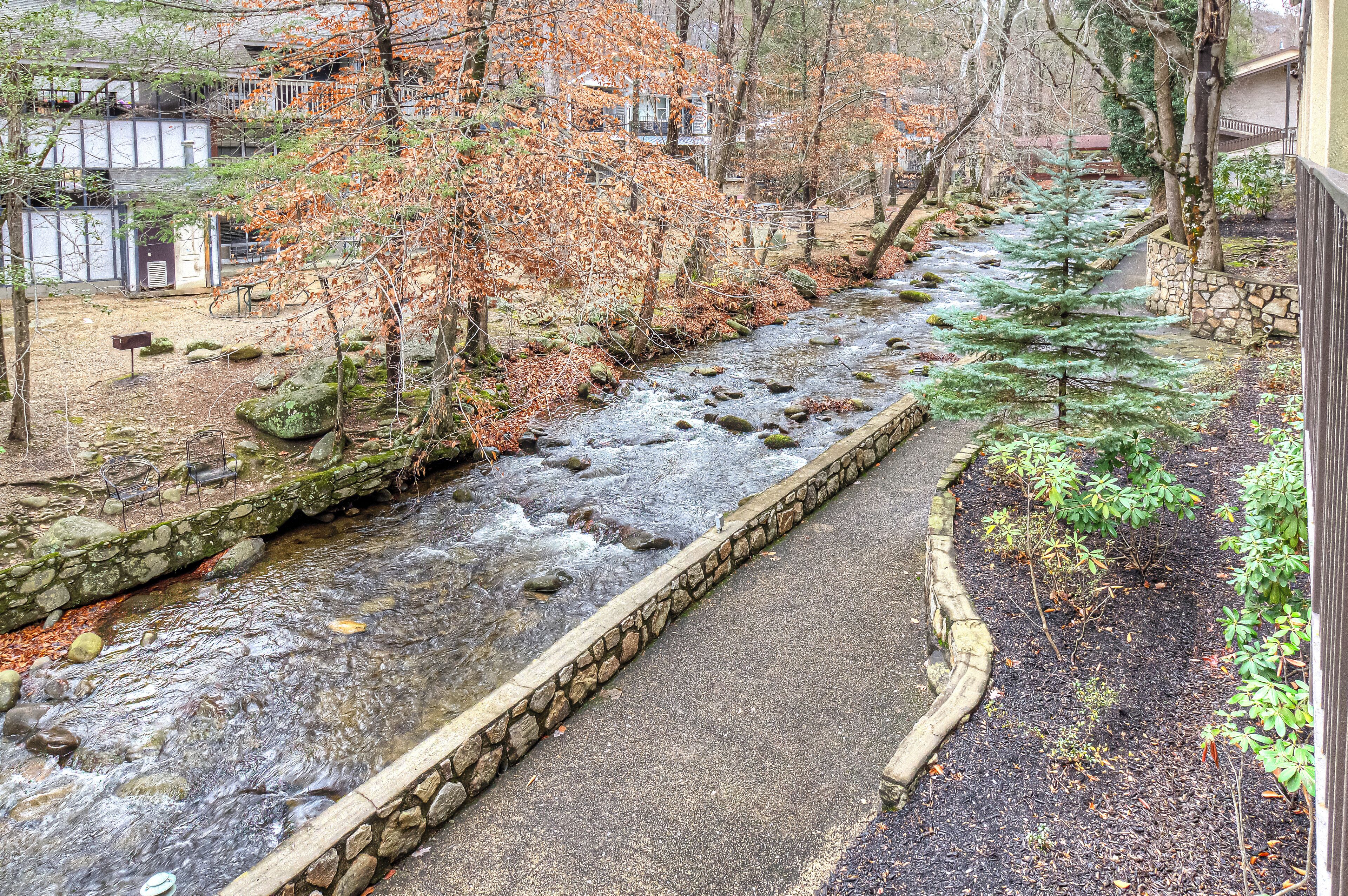 A stone-lined stream with a paved walkway, surrounded by trees and a stone wall.