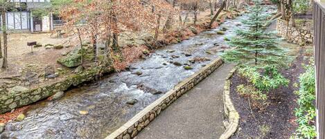 A stone-lined stream with a paved walkway, surrounded by trees and a stone wall.