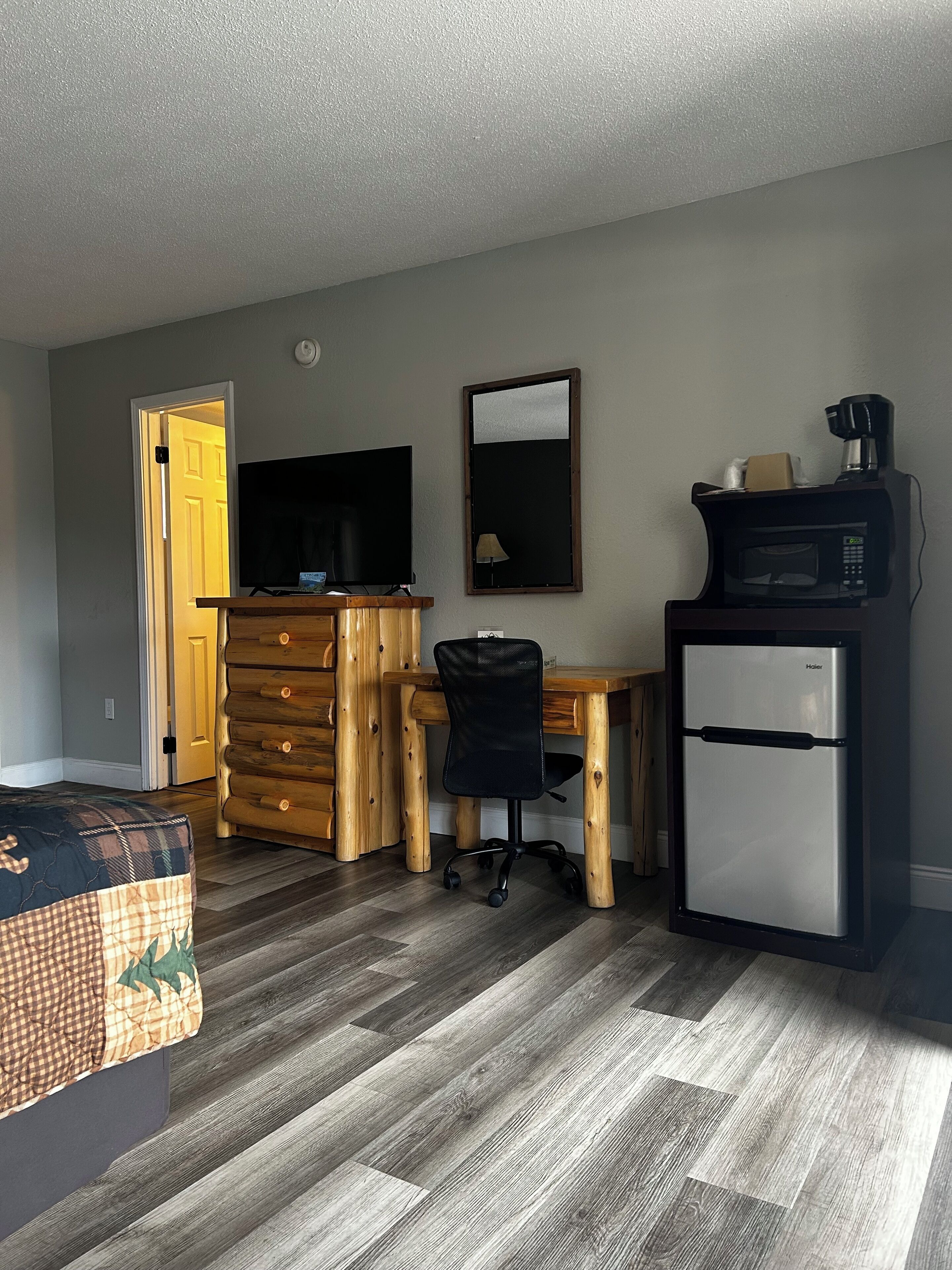 A compact room with a wooden desk, a black office chair, a television, a refrigerator, and a bed with a plaid bedspread.