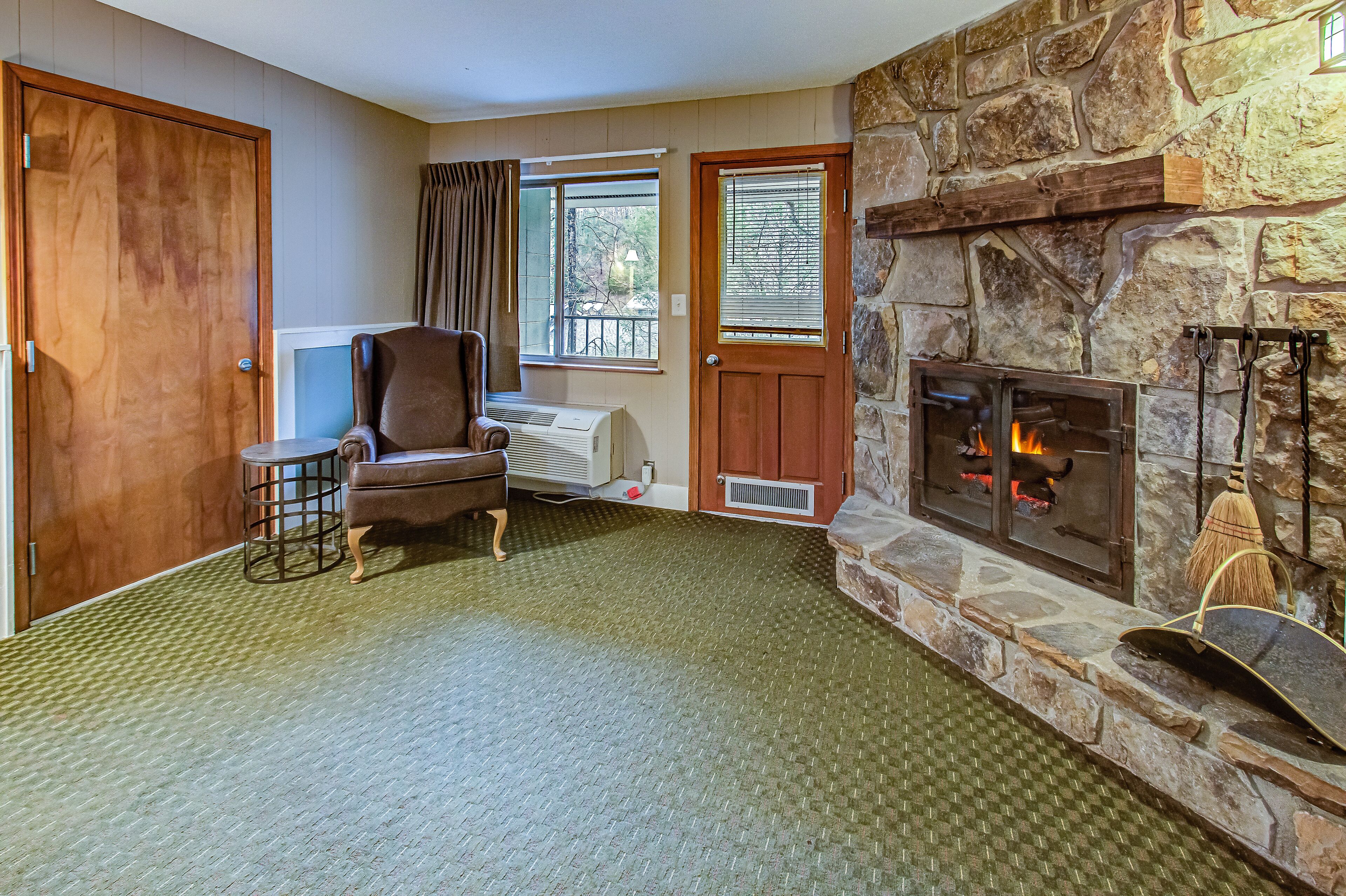 A cozy living room with a stone fireplace, a comfortable armchair, a small table, and a view through a window.