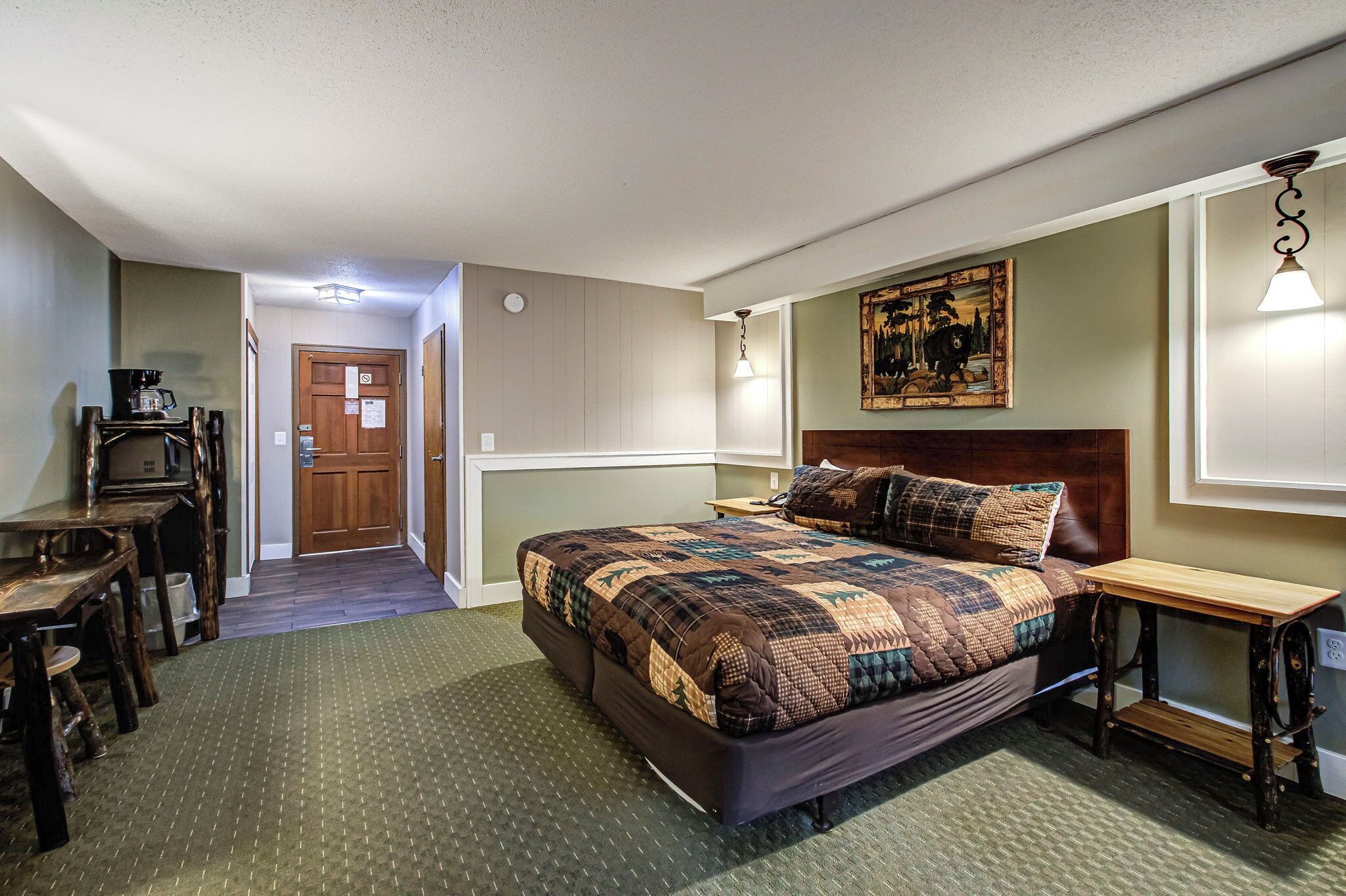 A bedroom with a large bed, a wooden nightstand, a desk, and a chair. There is a door leading to another room and a painting on the wall.