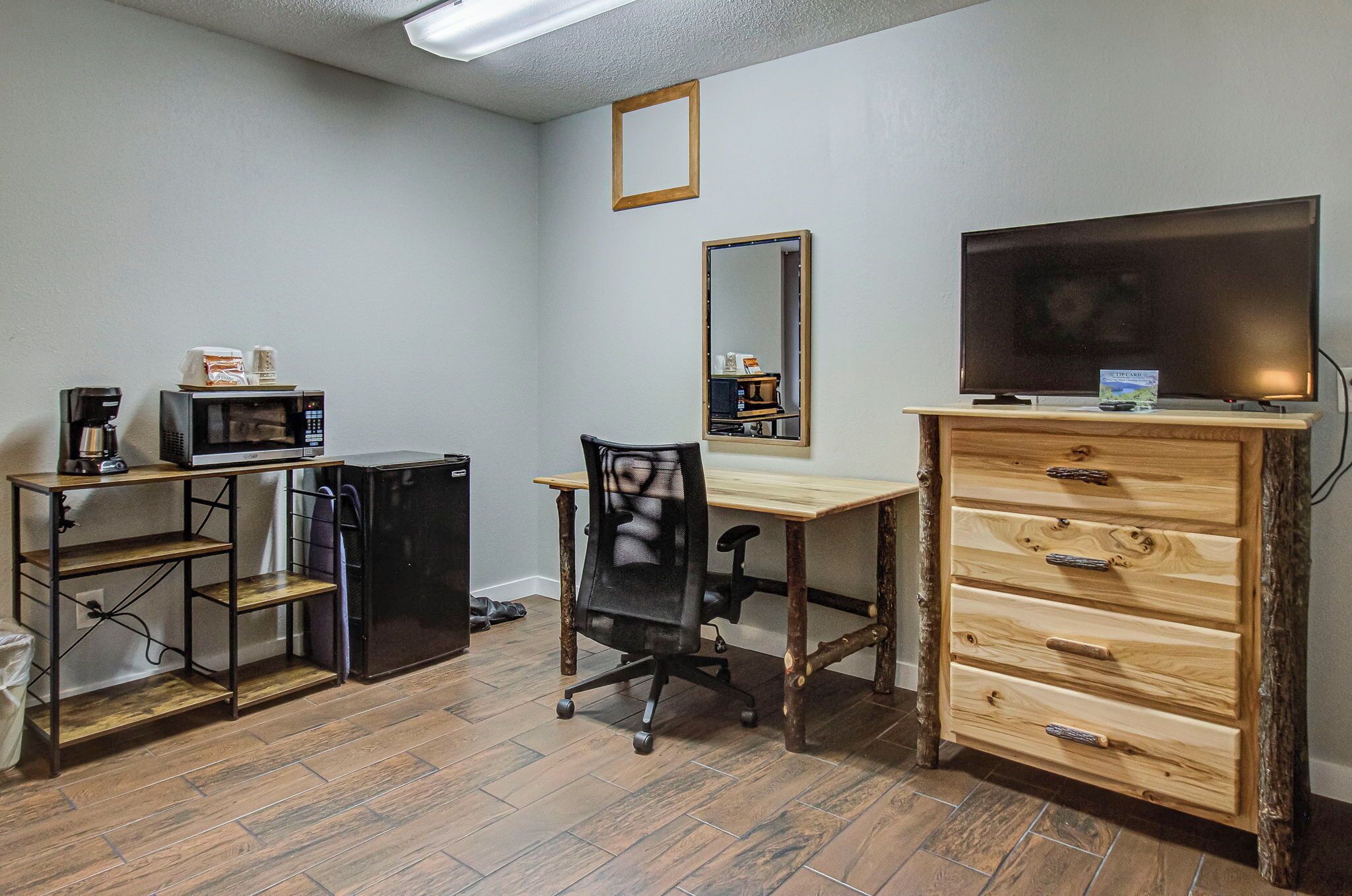 A small, furnished room with a wooden desk, a chair, a television, a microwave, and a dresser.