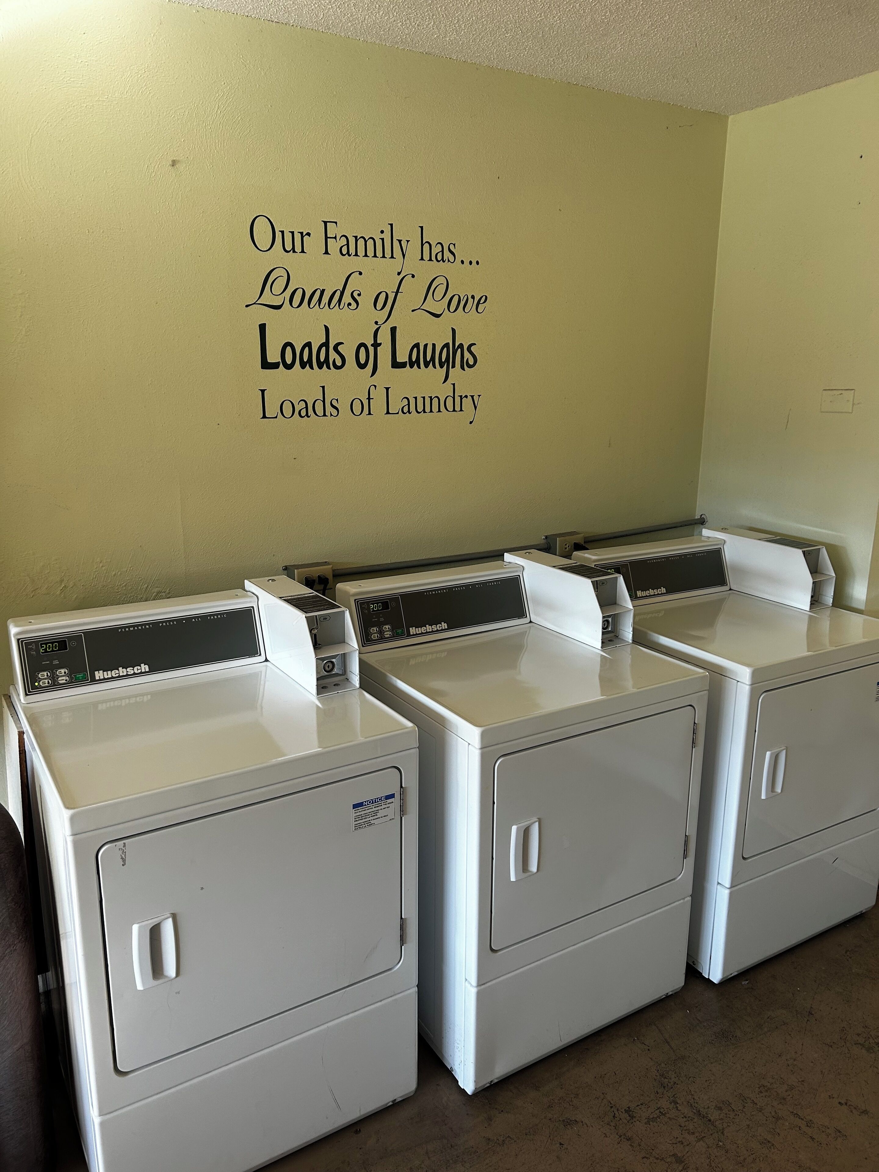 A laundry area with four white front-loading machines and a wall decal stating "Our Family has Loads of Love Loads of Laughs Loads of Laundry."