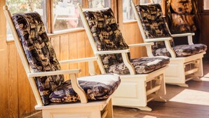 A row of wooden rocking chairs with patterned cushions, positioned in a sunlit room with large windows overlooking a street. - Brookside Lodge (Gatlinburg)