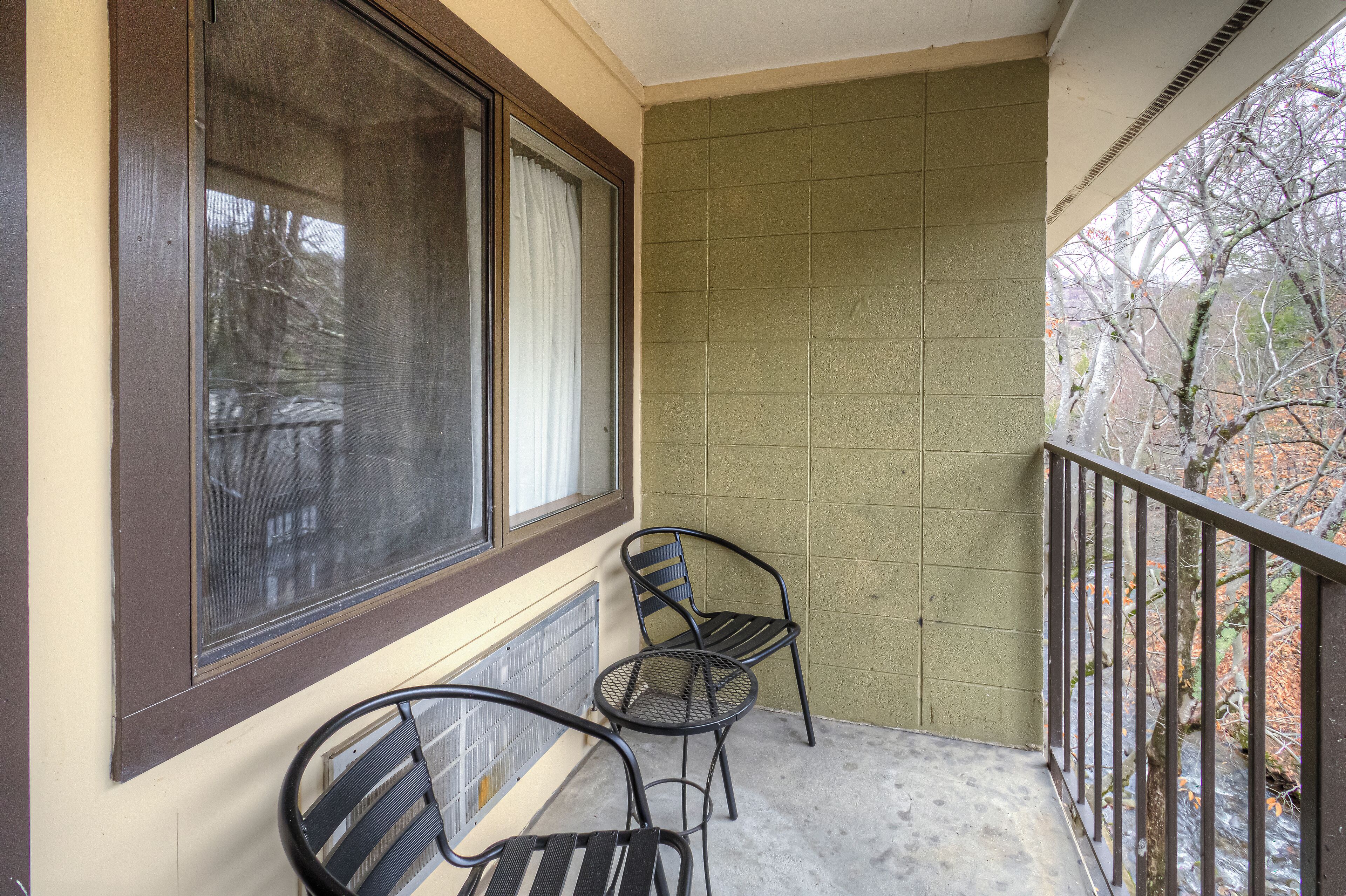 Un balcon avec deux chaises en métal, une fenêtre avec un rideau et une vue sur les arbres.