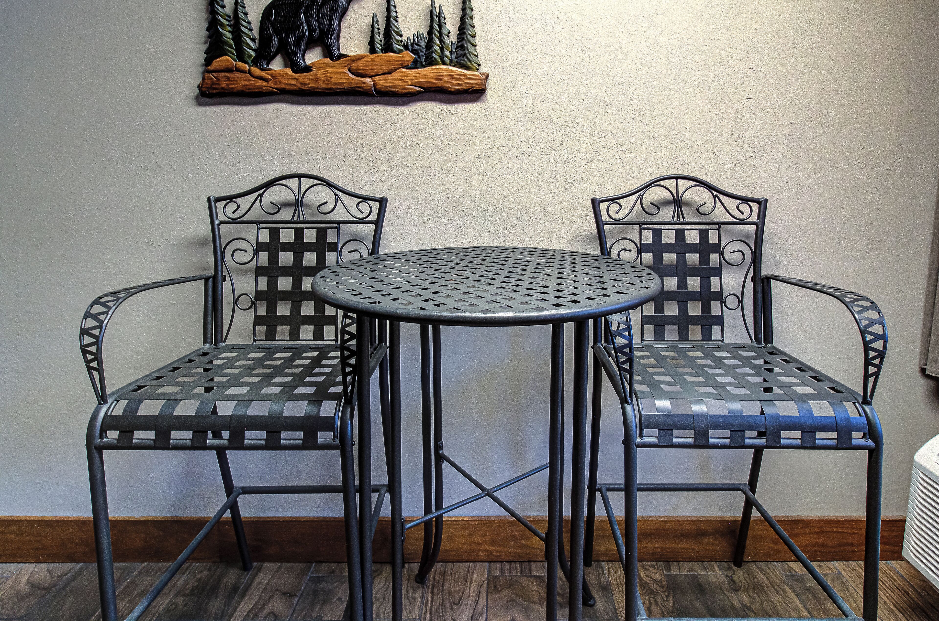 A small, round metal table with two matching chairs, a wooden shelf with a bear decoration, and a white wall.