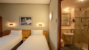 Habitación de hotel con dos camas, baño con ducha de vidrio y lavamanos montado en la pared.