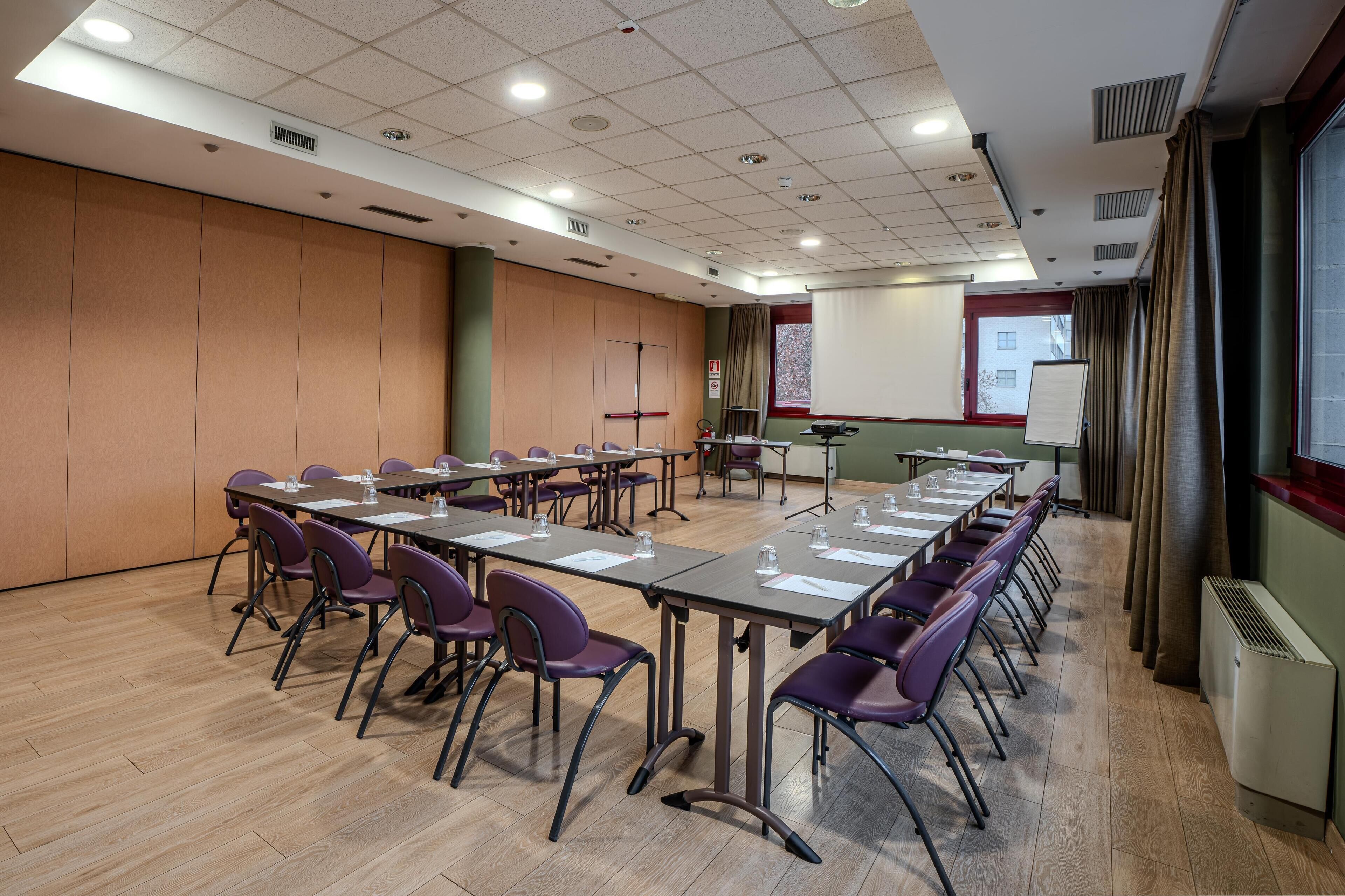 Une salle de conférence équipée de longues tables et de chaises, d’un écran de projection et d’un tableau blanc.