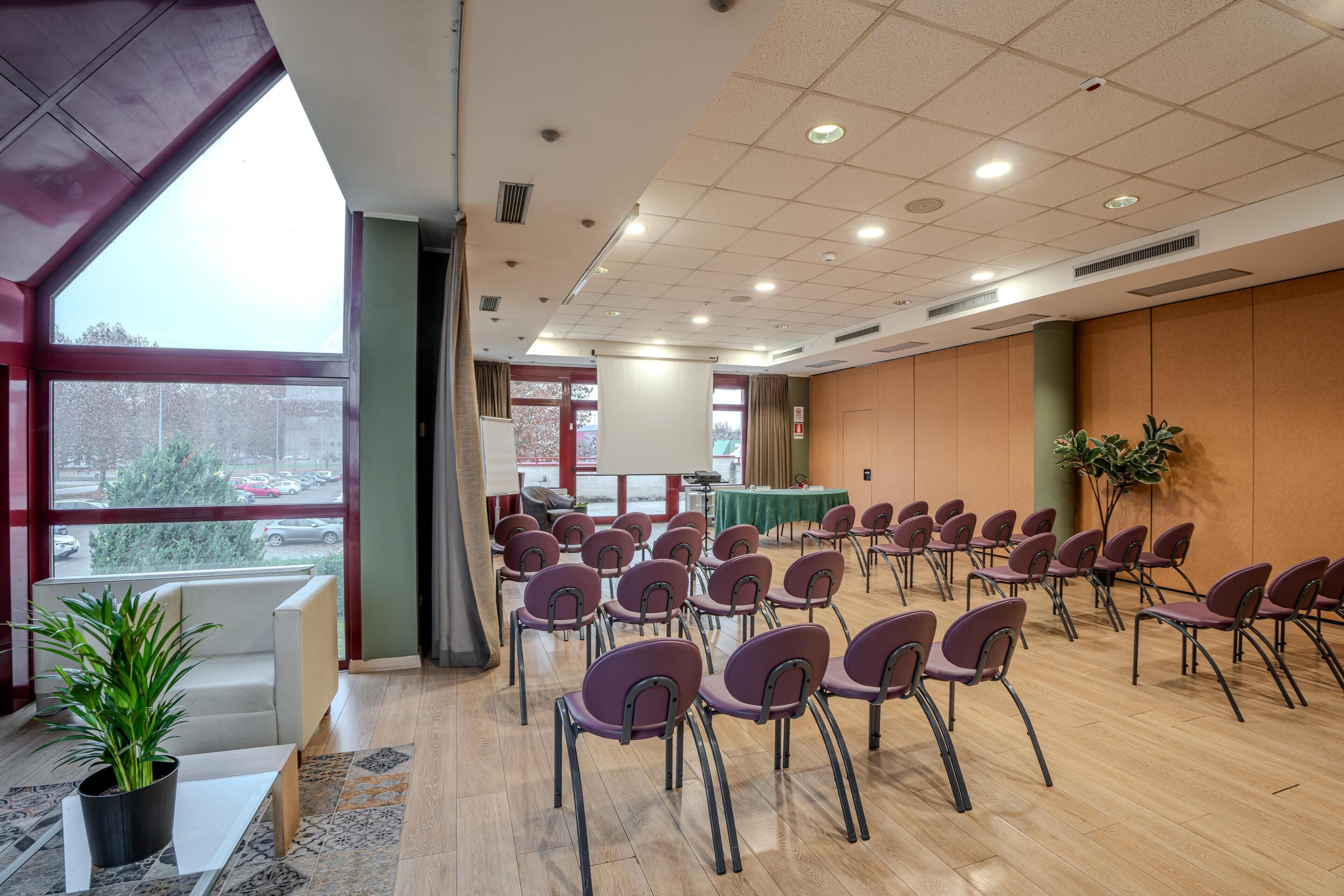 Uma sala de conferências com fileiras de cadeiras, um palco e uma grande janela com vista para um estacionamento.