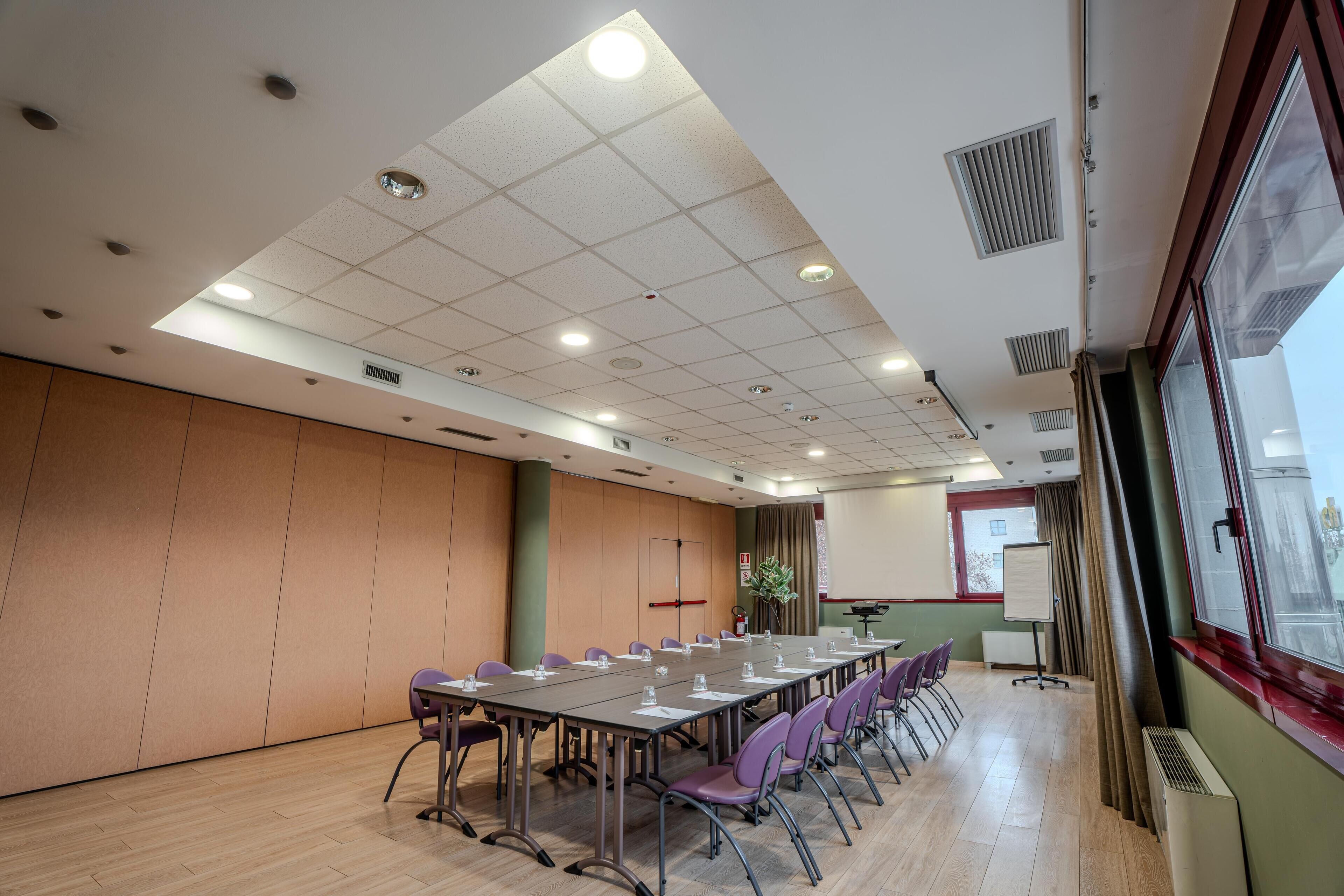 Un salón de conferencias con una larga mesa, sillas moradas, una pantalla de proyector y un ventanal con cortinas.