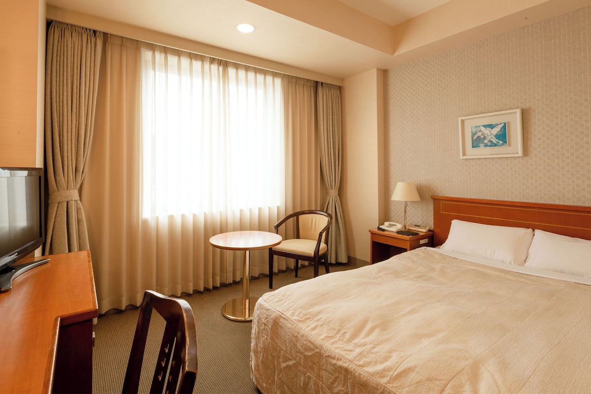 A hotel room with a large bed, a desk with a television, a chair, a small table, and a lamp.