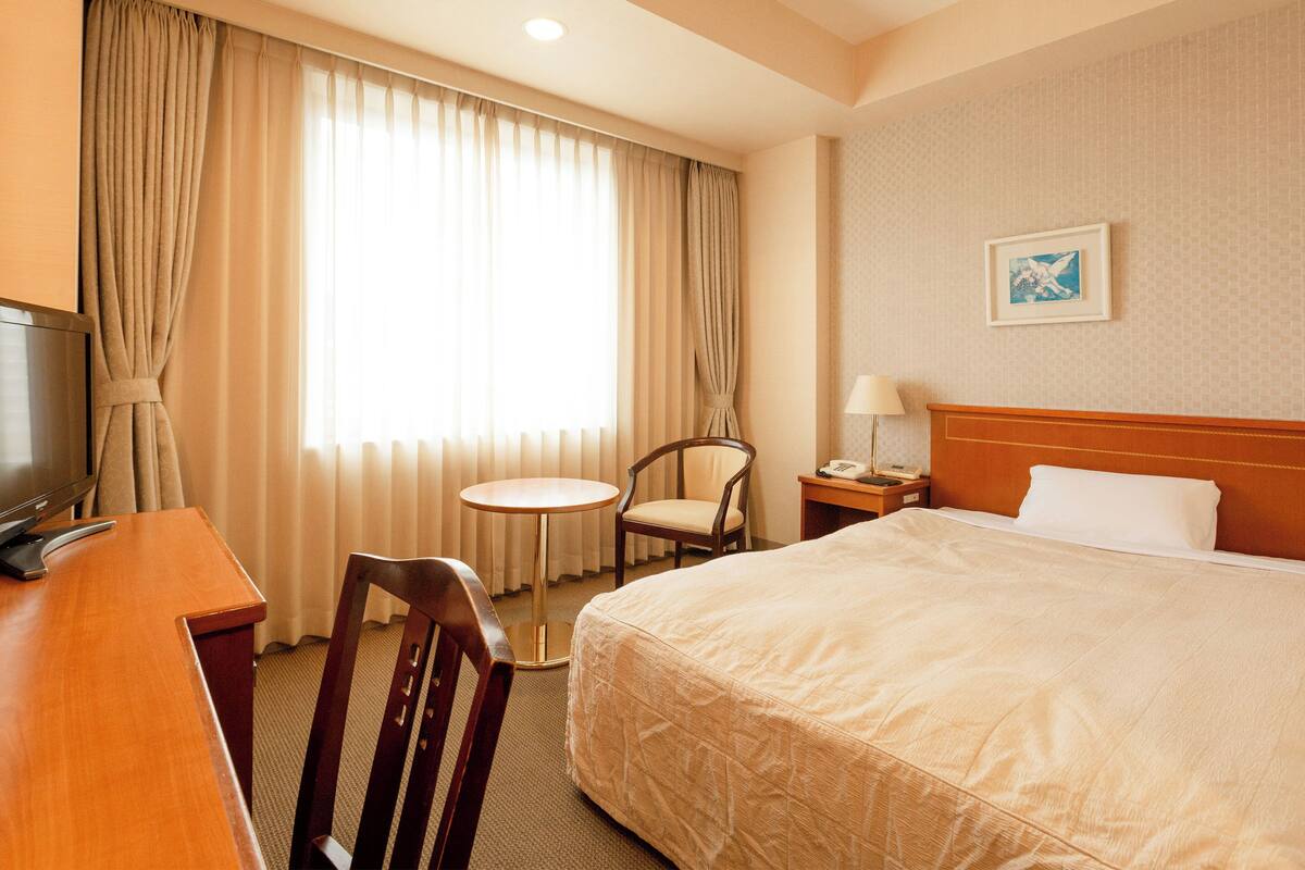 A hotel room with a bed, a desk with a television, a chair, a small table, and a nightstand with a lamp.