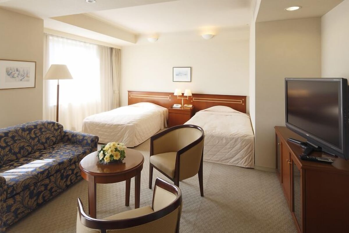 A hotel room with a sofa, two single beds, a chair, a small table with flowers, a TV, and a lamp.