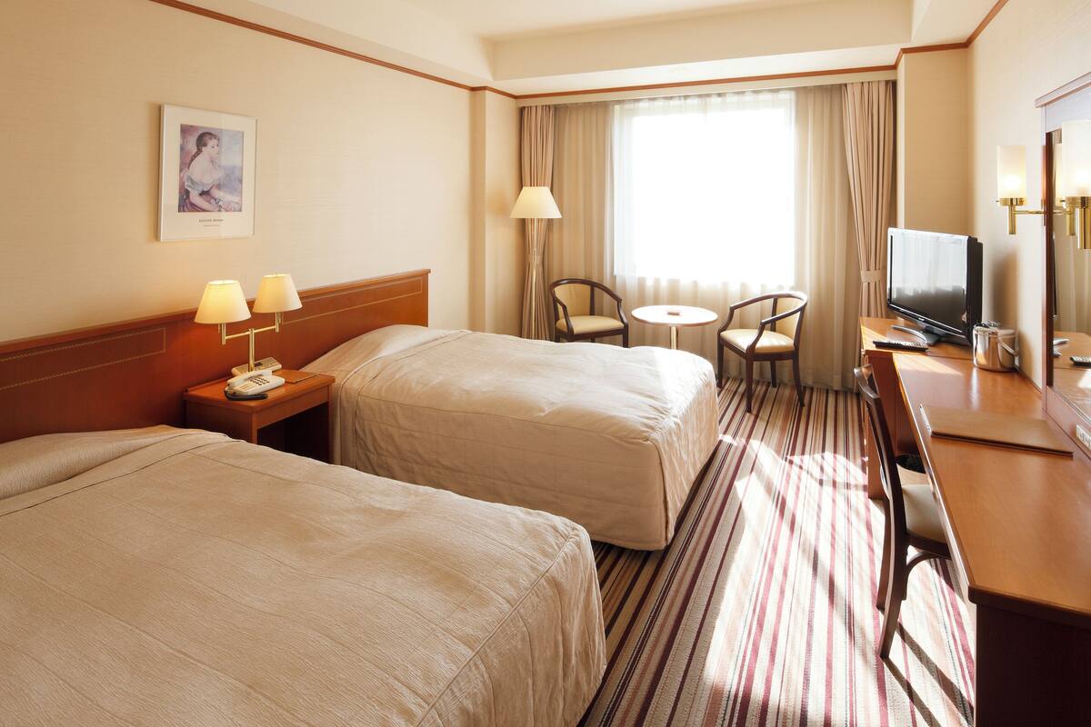A hotel room with two beds, a desk with a TV, a small table with two chairs, and a window with curtains.