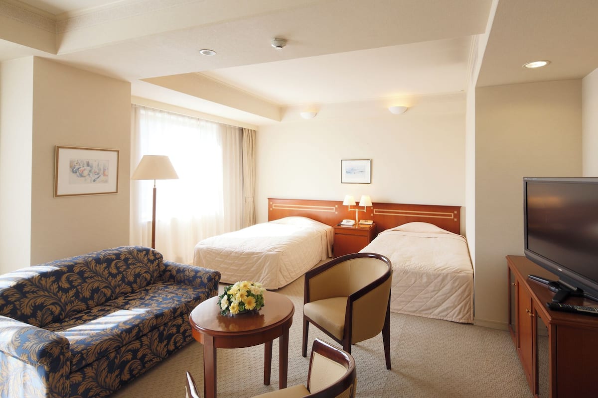A hotel room with two beds, a sofa, a small table with a vase, a TV, and a lamp.