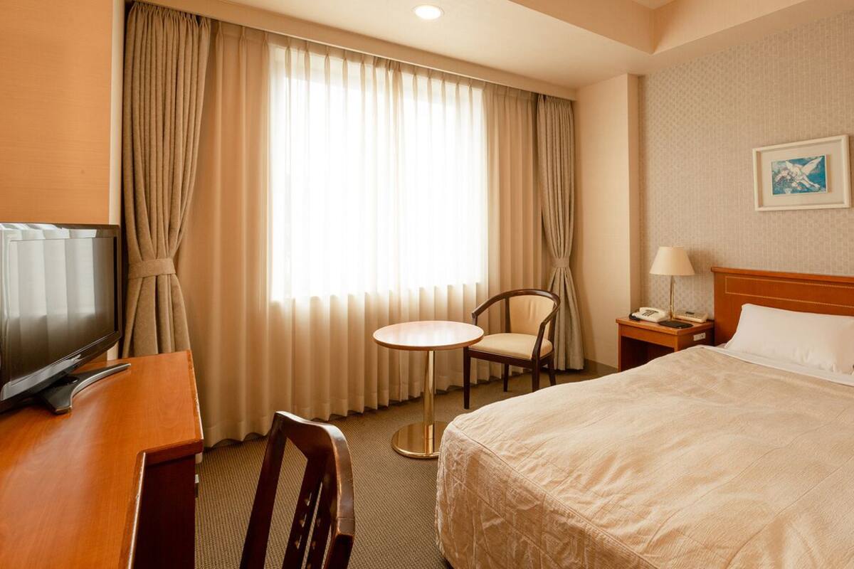A hotel room with a bed, a desk with a TV, a chair, a small table, a lamp, and a painting on the wall.