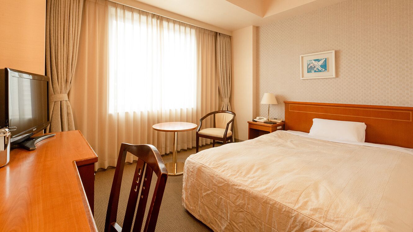 A hotel room with a bed, a desk with a television, a chair, a small table, and a nightstand with a lamp.