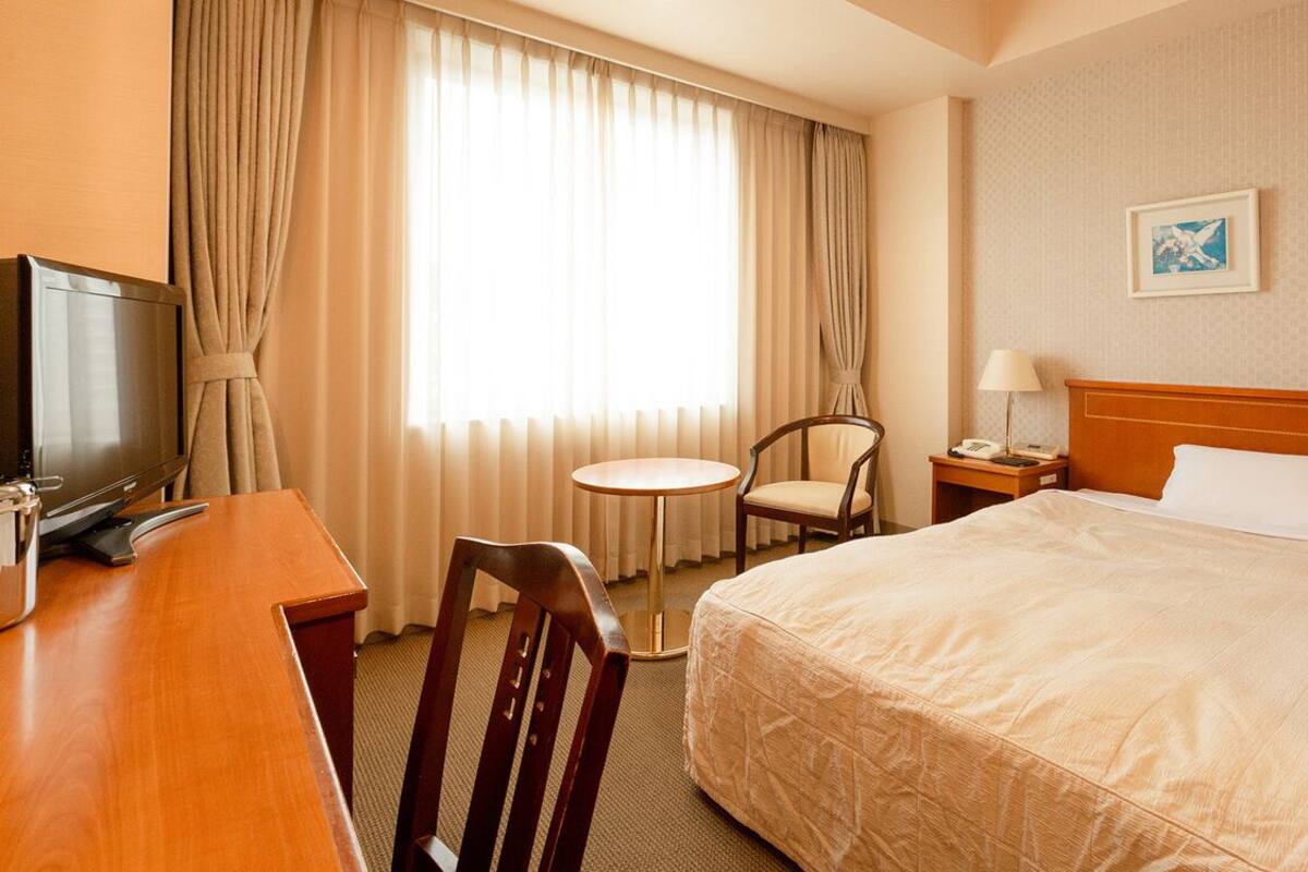 A hotel room with a bed, a desk with a television, a chair, a small table, and a nightstand with a lamp.