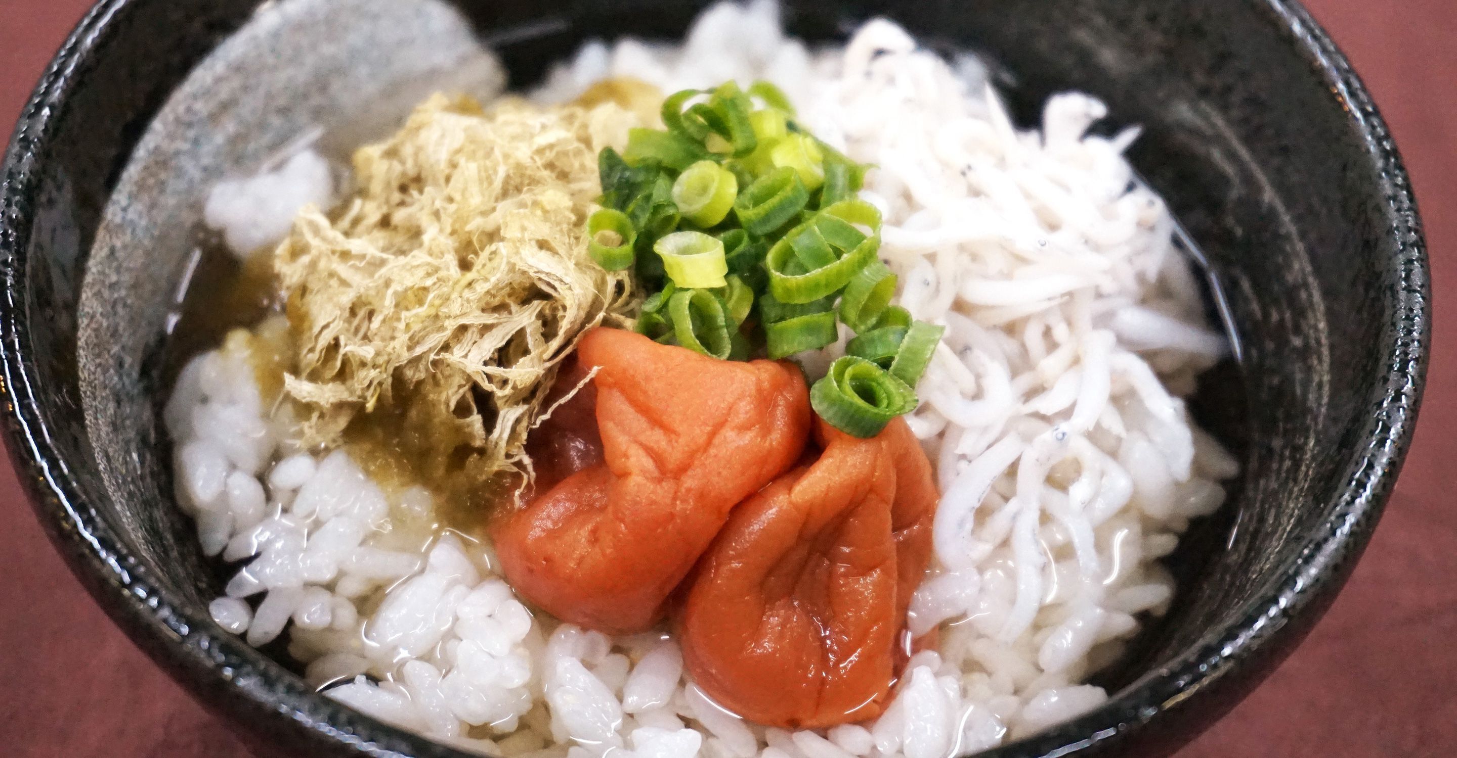 A bowl of white rice with toppings including shredded seaweed, sliced green onions, and a piece of carrot.