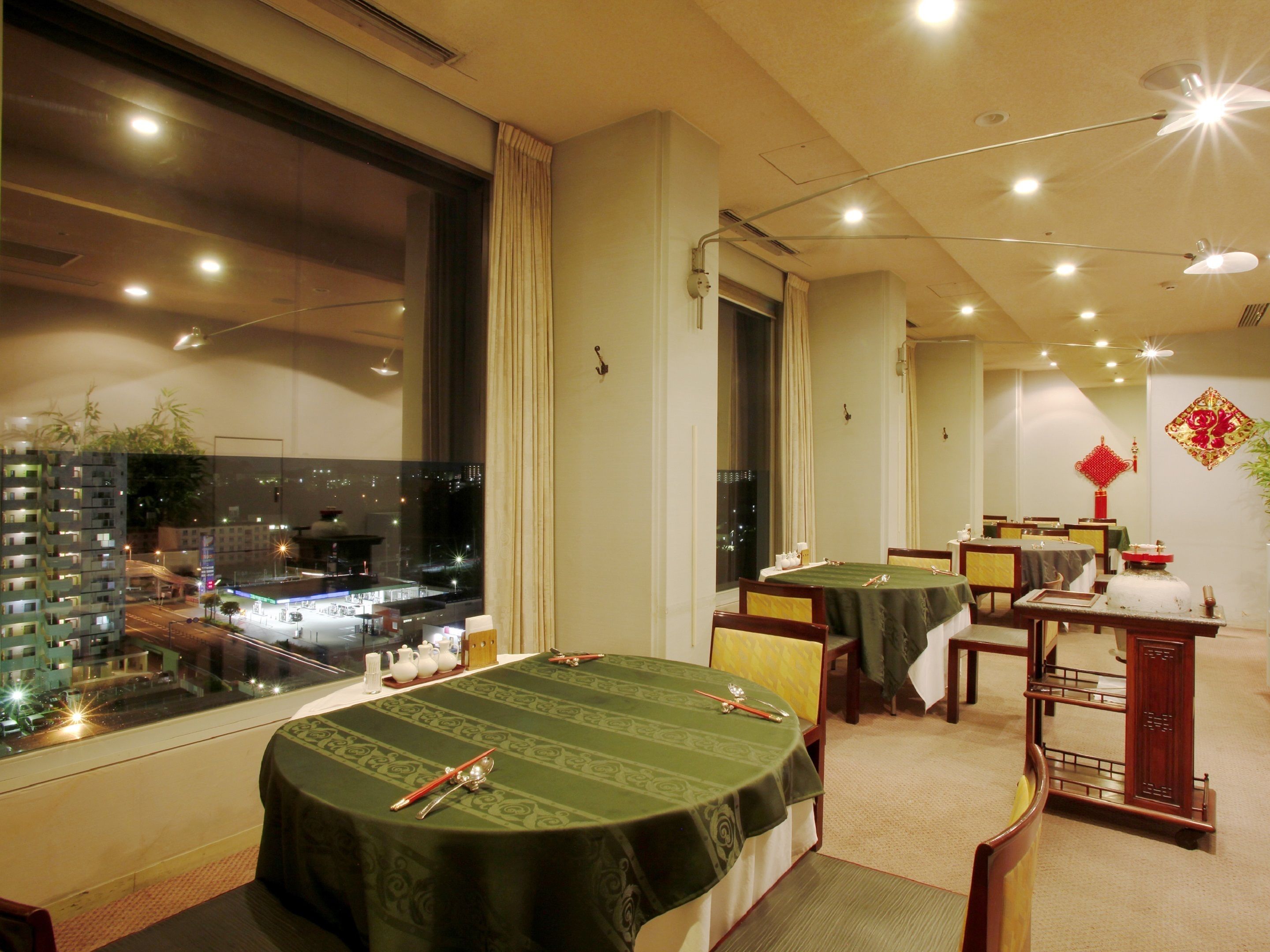 A dining area with round tables covered in green tablecloths, a view of a cityscape through large windows, and traditional decor including a red Chinese knot on the wall.