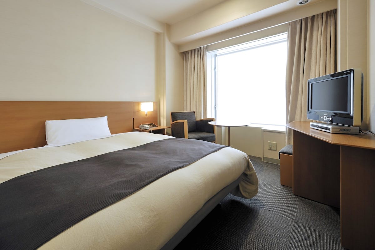 A hotel room with a large bed, a desk with a television, a chair, and a window with curtains.