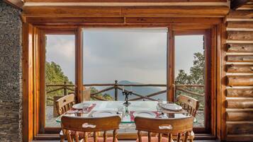 A wooden cabin with a dining table set for two, large windows, and a view of the mountains.