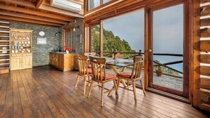 A wooden interior with a dining area, stone wall, and large windows overlooking a scenic view. - Rokeby Manor (Mussoorie)