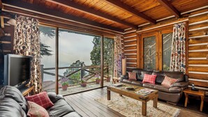 A log cabin interior with a wooden ceiling, a leather sofa, a coffee table, and large windows with curtains. - Rokeby Manor (Mussoorie)