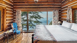 A log cabin room with a large bed, a chair, a table, and a view of mountains. - Rokeby Manor (Mussoorie)
